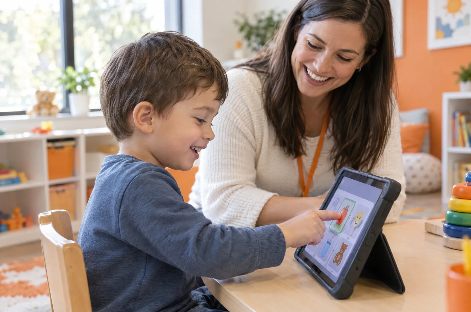 A boy taps a picture button on a communication tablet as his therapist smiles