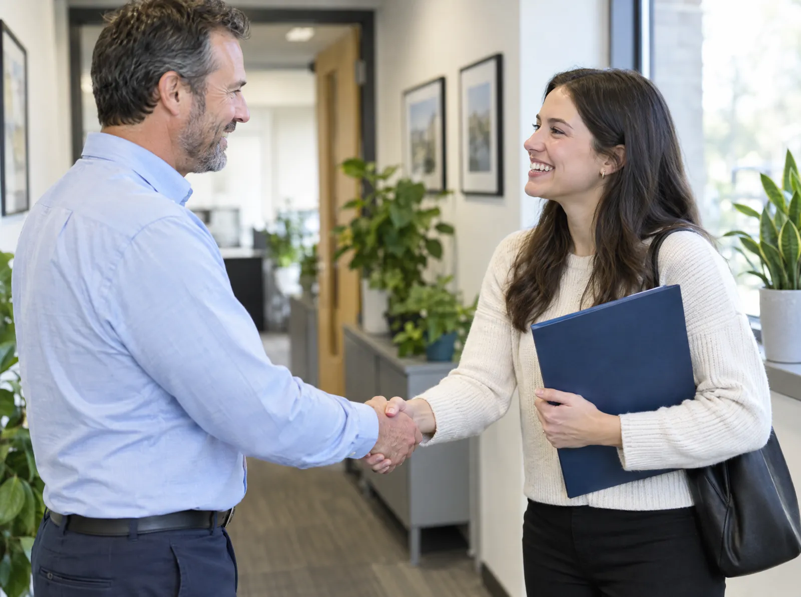 An office manager shakes hands with a smiling new hire holding a welcome folder