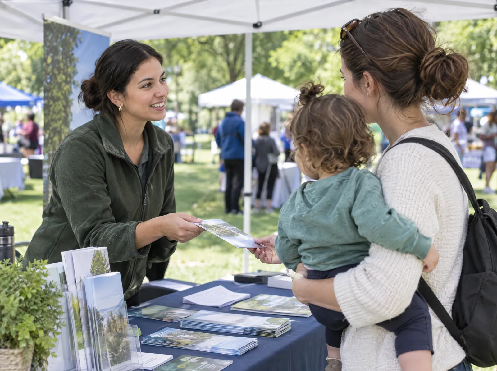 A representative hands a brochure to a mother holding her toddler under a canopy tent