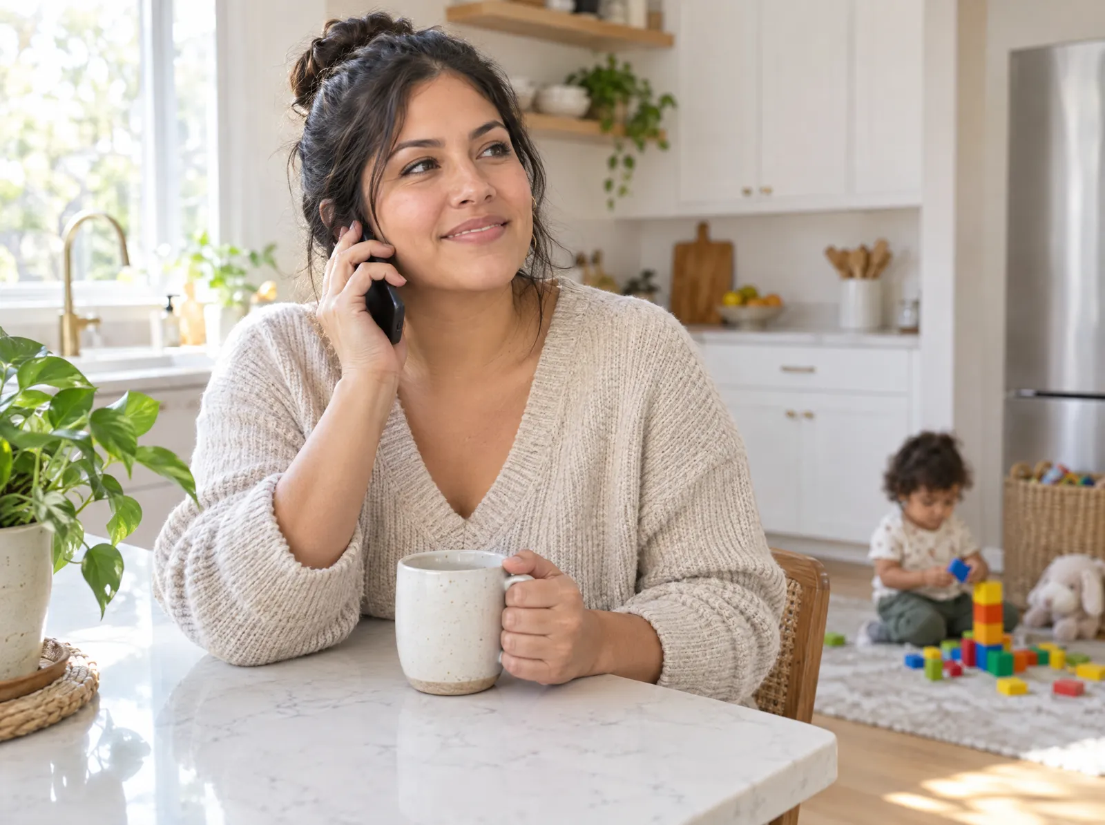 A smiling mother talks on the phone in her kitchen while her toddler plays nearby